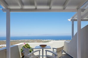 A balcony with a table and two chairs, overlooking a coastal landscape with the sea and distant hills. - Anchor Suites (Santorini)