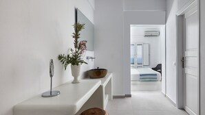 A modern bathroom with a white vanity, a wooden stool, a mirror, and a plant. - Anchor Suites (Santorini)