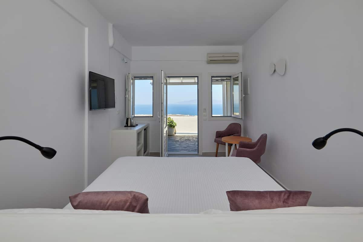 Habitación de hotel con cama, escritorio, silla y televisión. Hay dos ventanas con vista al mar.