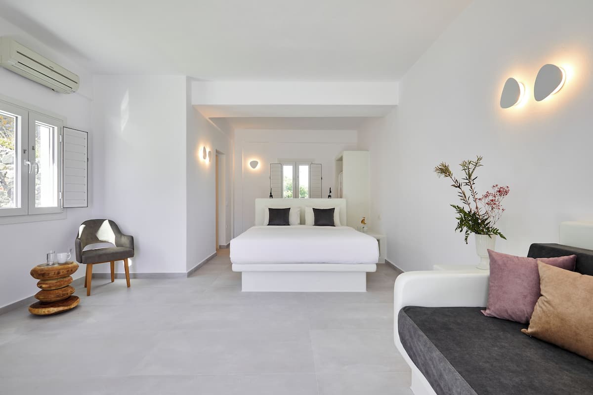 Un dormitorio moderno con una cama grande, un sofá gris, una mesita de noche con un jarrón y una silla.