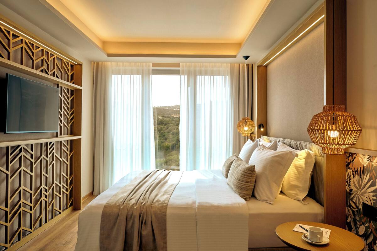 Habitación de hotel con una cama grande, un televisor instalado en un panel de madera, una ventana con cortinas y una mesa con una taza.