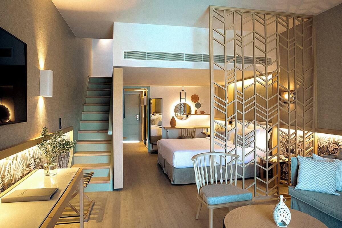 Una habitación de hotel moderna con cama tipo loft, una zona de comedor con mesa y sillas, y una sala de estar con sofá y objetos decorativos.