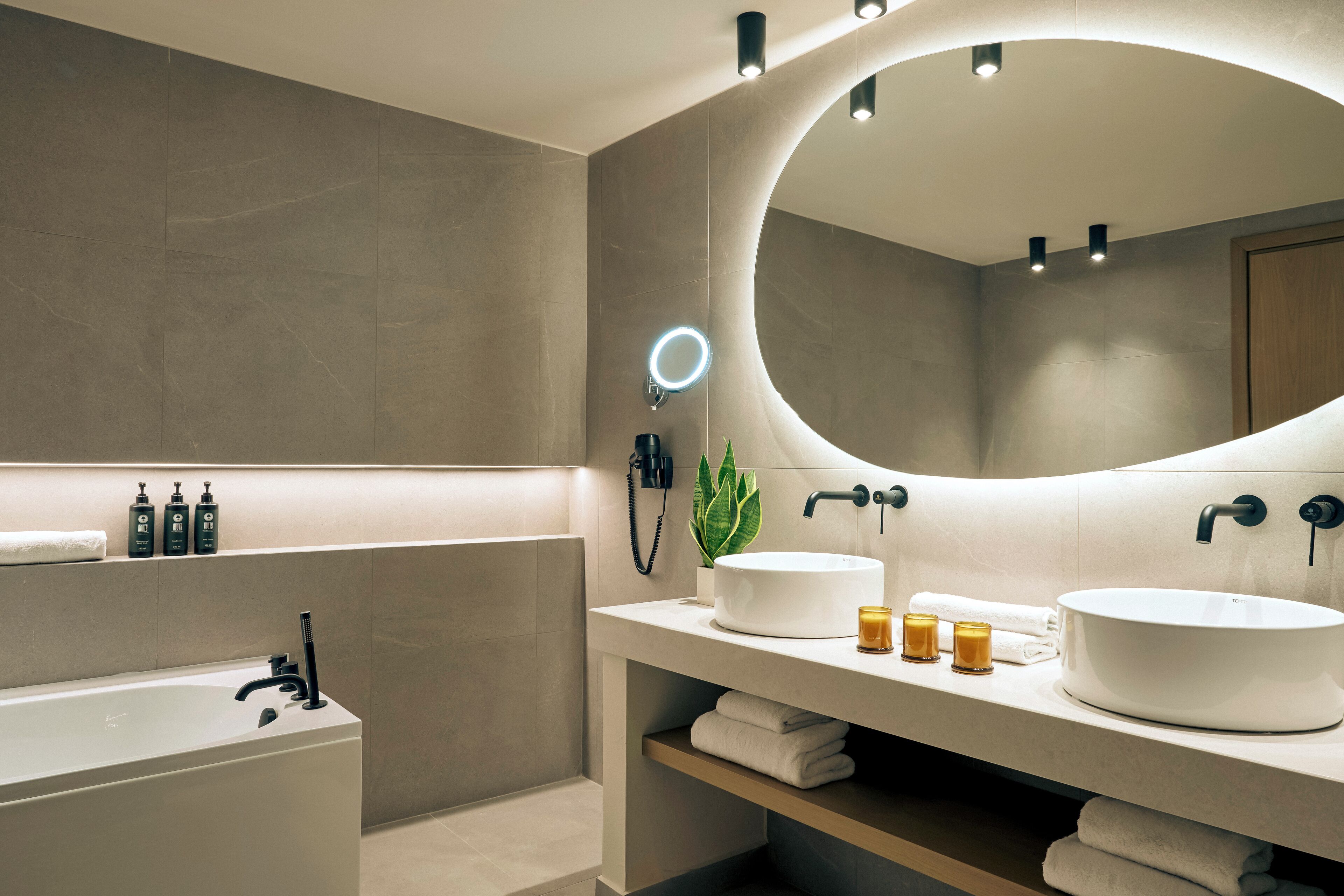 Ein modernes Badezimmer mit einer Badewanne, einem großzügigen Waschbecken und einem Spiegel mit runder Beleuchtung. Auf einem Regal liegen Pflegeprodukte und auf der Ablage steht eine Pflanze.