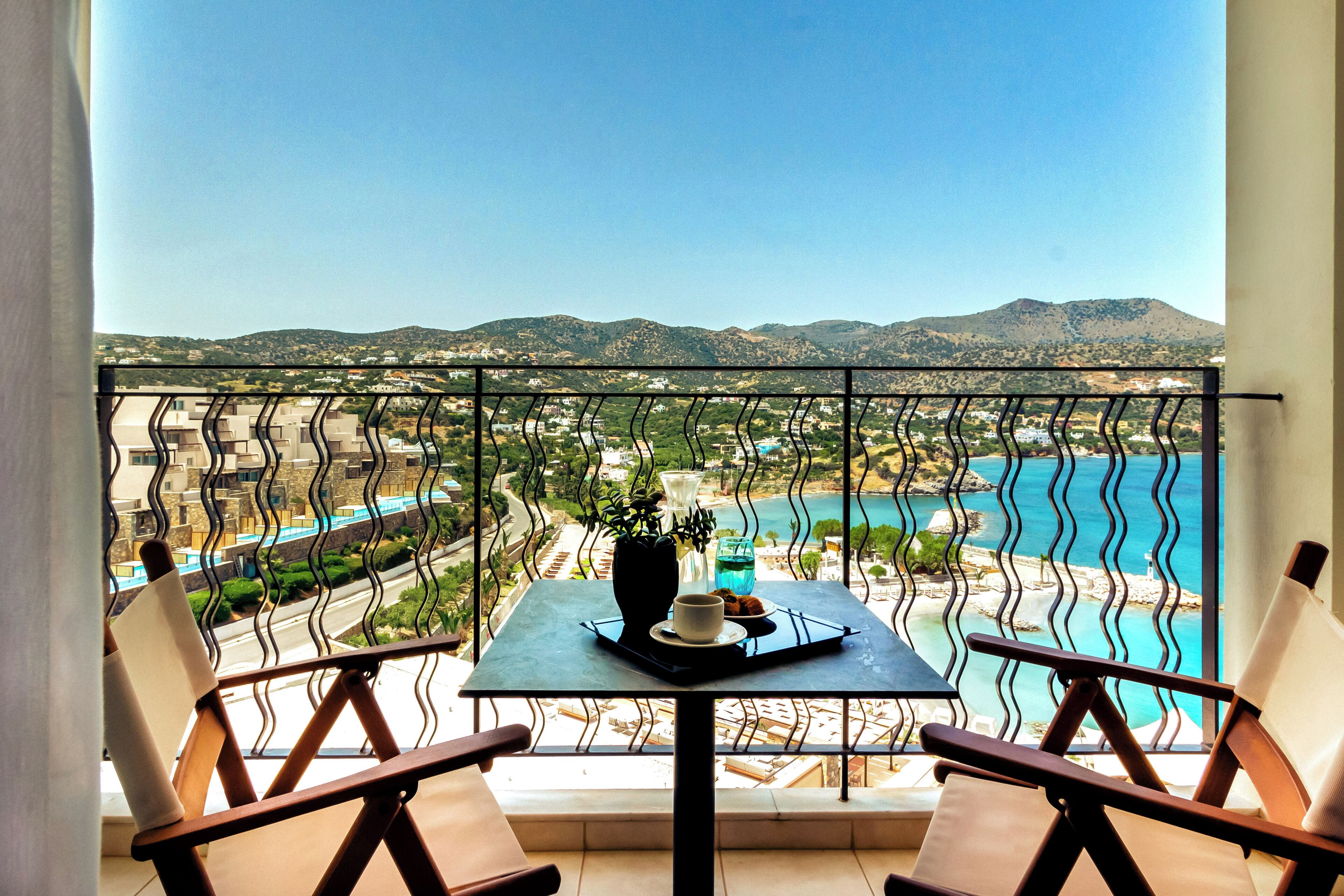 A balcony with a table, two chairs, and a view of a coastal area with mountains in the background.