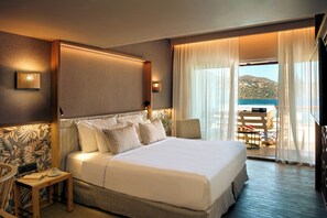 Ein Hotelzimmer mit einem großen Bett, einem Nachttisch mit Tasse und Buch, einem Sessel und Meerblick durch das Fenster.