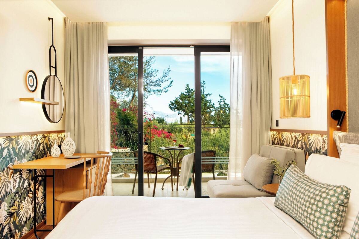 Habitación de hotel con cama, sofá, mesa de comedor con sillas y balcón con vistas a árboles y flores.