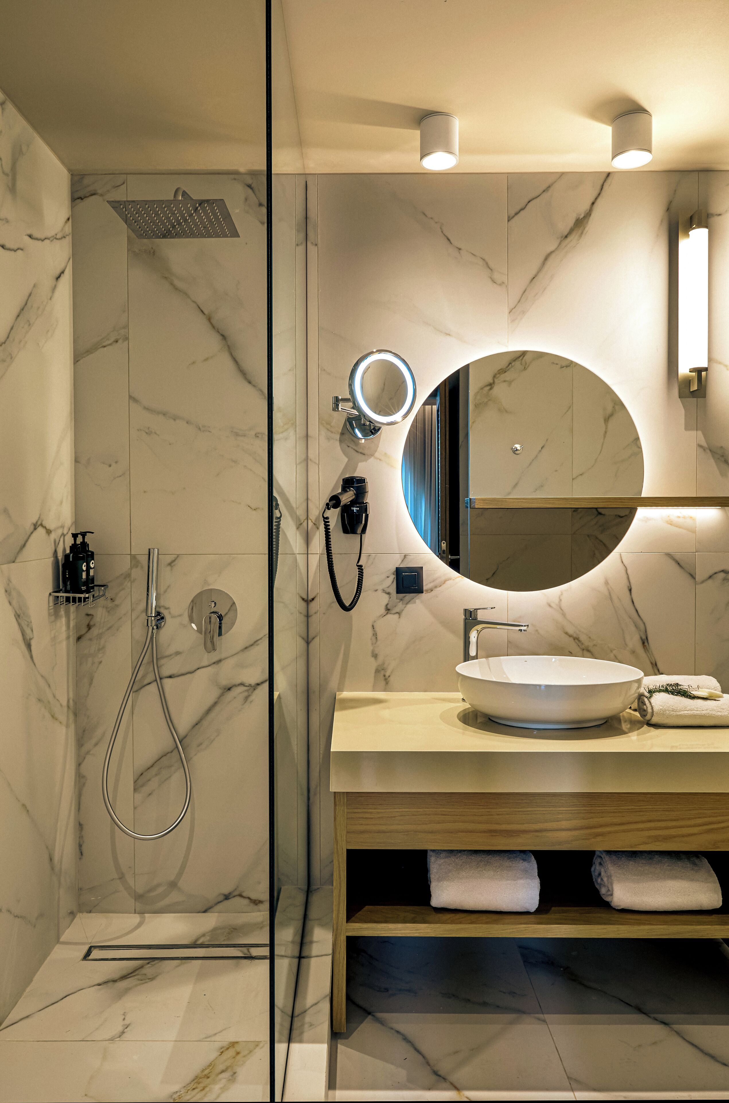 Un baño moderno con ducha de vidrio, espejo redondo, lavamanos blanco y estantes de madera con toallas.