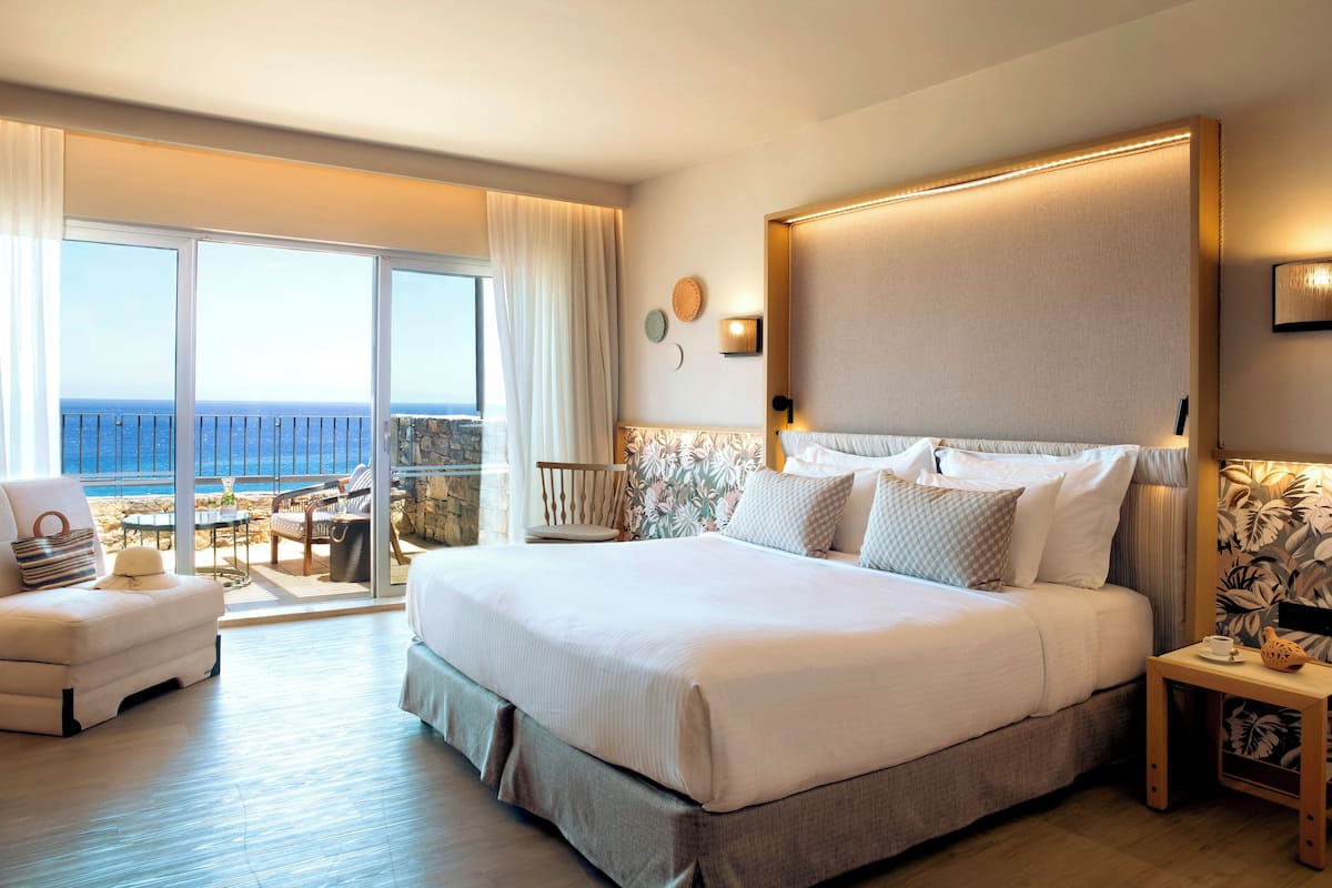 Un dormitorio espacioso con una cama grande, un rincón de estar con una silla y una mesita, y un balcón con vistas al mar.