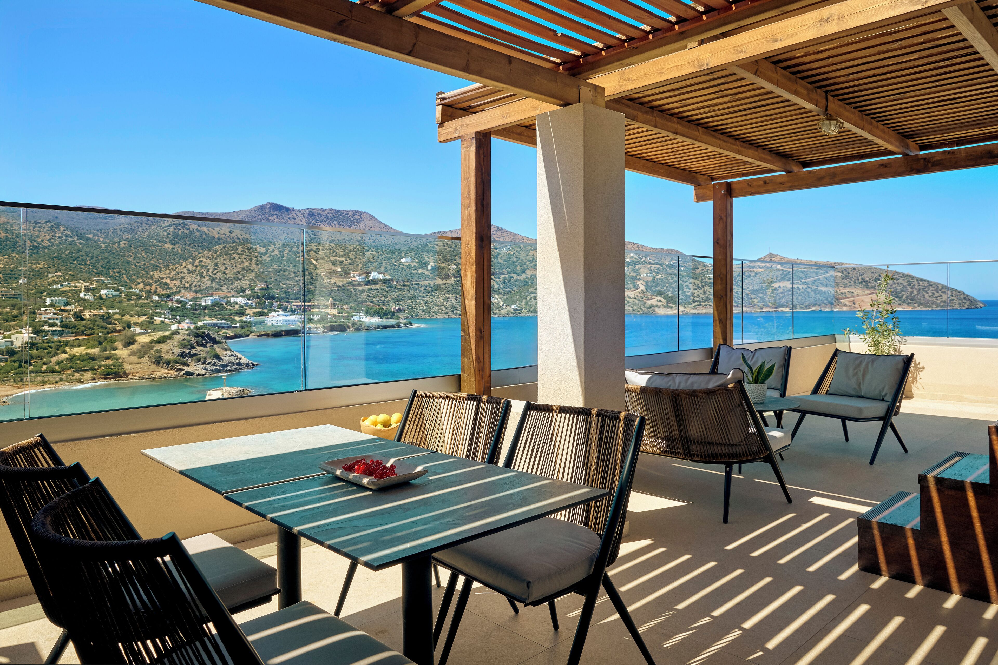 Terraço com mesa e cadeiras, dando vista para uma paisagem costeira com montanhas e um céu azul e límpido.