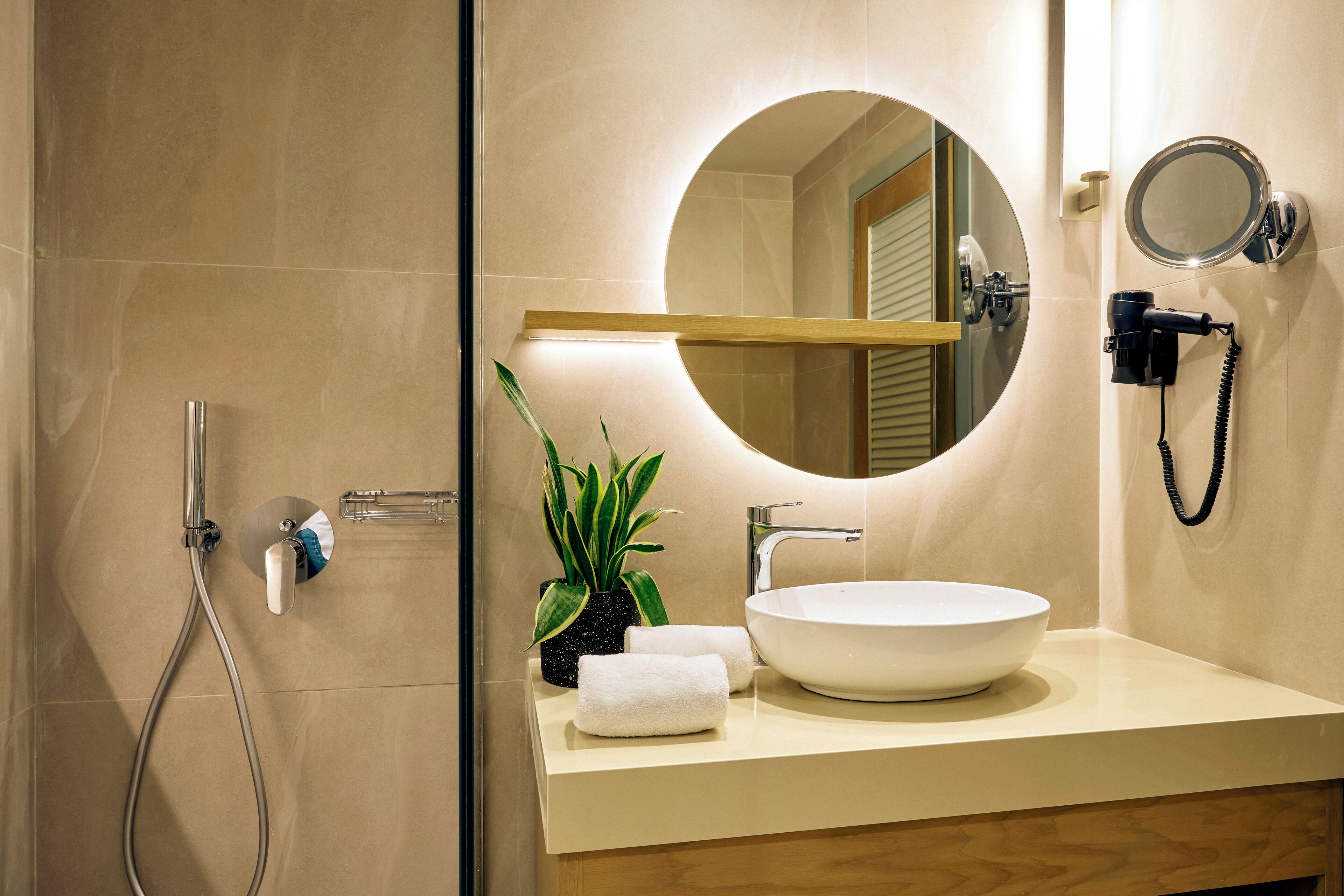 A modern bathroom with a round mirror, a sink, a handheld showerhead, and a potted plant.