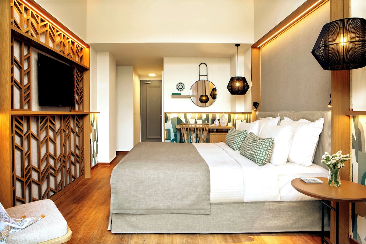 Una habitación de hotel moderna con una cama grande, un televisor montado en un panel de madera, un reloj de pared redondo y una mesita de noche con un jarrón de flores.
