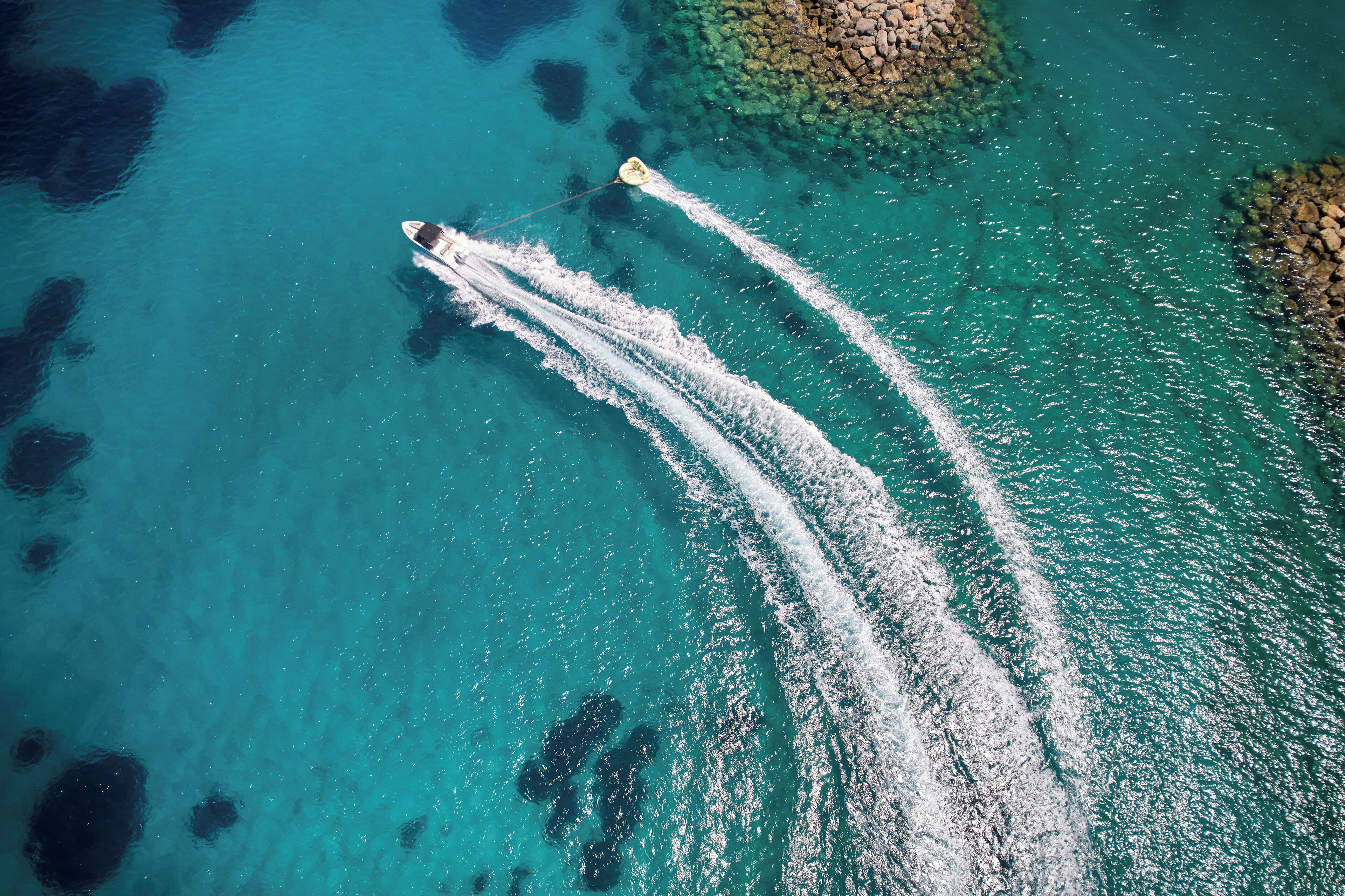 An aerial view of a boat creating wake patterns in clear turquoise water with rocky formations.