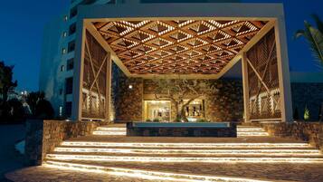 A modern building with a geometric wooden ceiling structure, stone walls, and a well-lit entrance with steps leading up to a seating area.