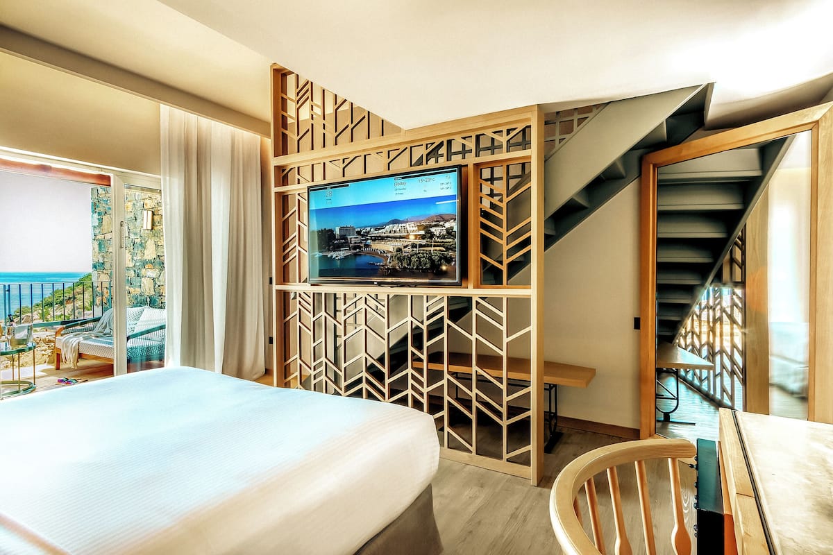 Habitación de hotel con una cama, un televisor montado en una separadora de madera, una silla y vista al mar a través de un ventanal.