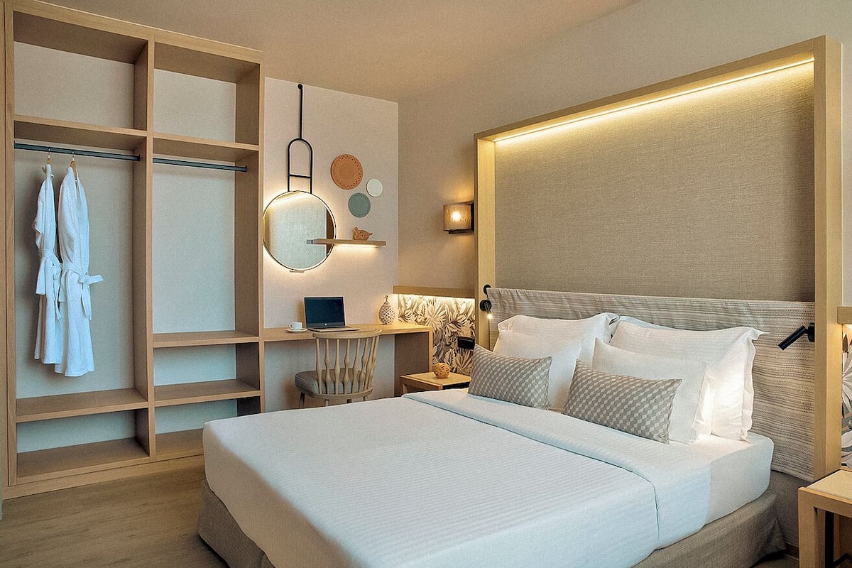 Una habitación de hotel moderna con una cama grande, un armario empotrado, un escritorio con una computadora portátil y un espejo montado en la pared.