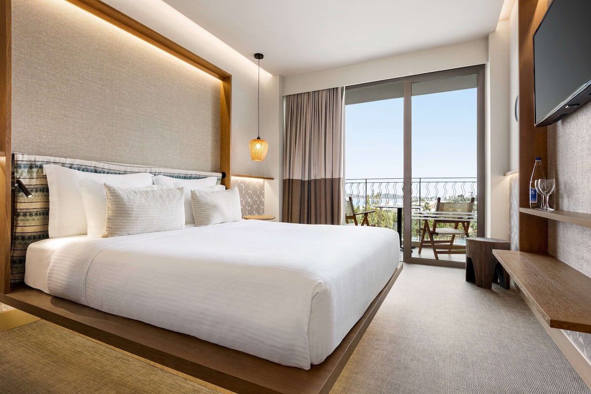 Habitación de hotel con una cama grande, televisión, balcón con mesa y sillas, y vista al cielo.