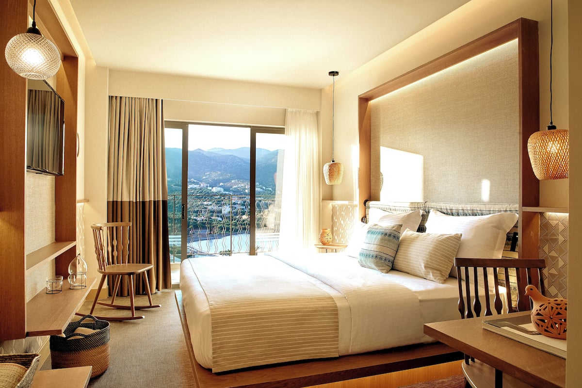 Habitación de hotel con una cama grande, cabecera de madera, escritorio con silla, vista a las montañas y un balcón.