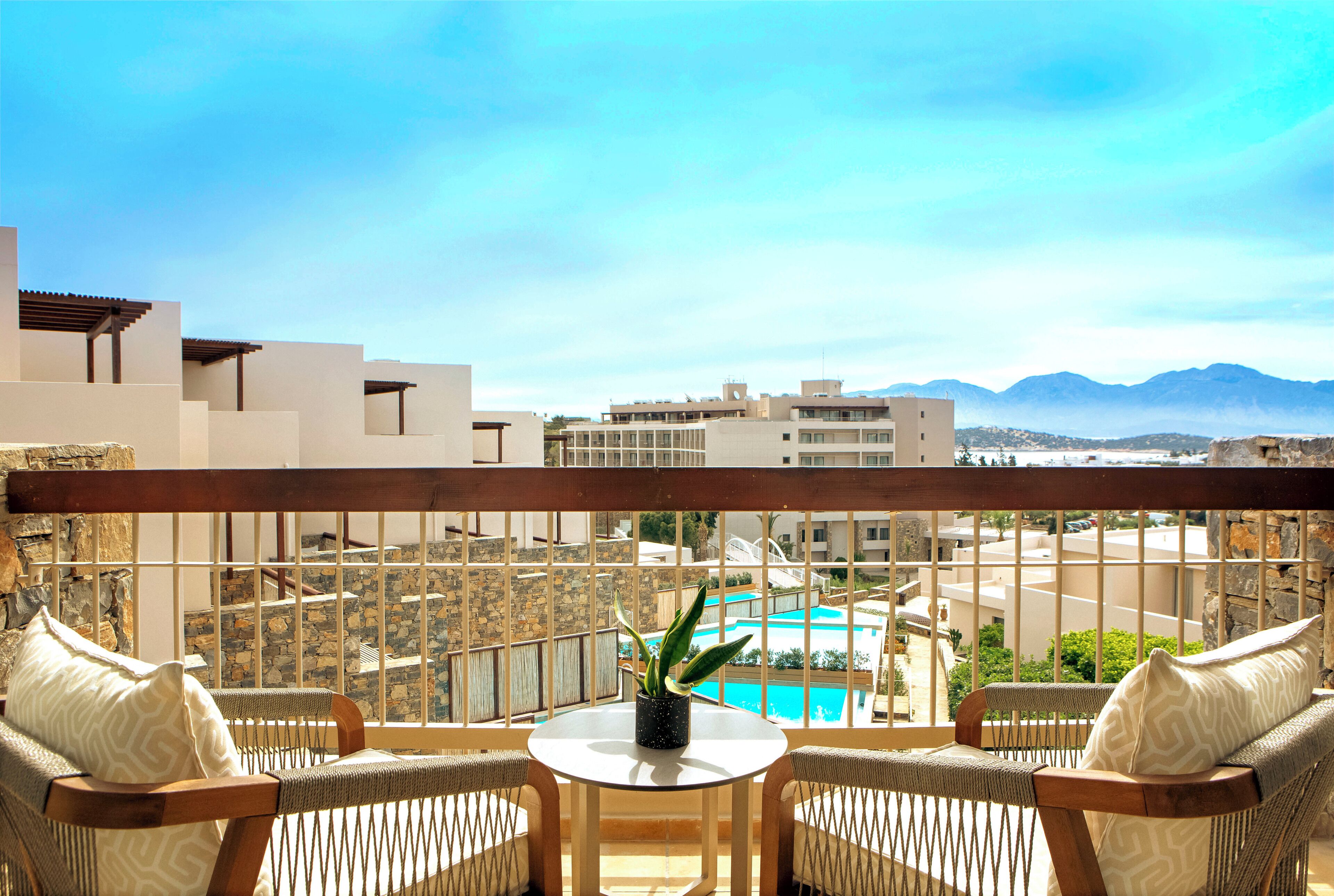 A balcony with wicker furniture, a small table, and a view of a pool and buildings in the distance.