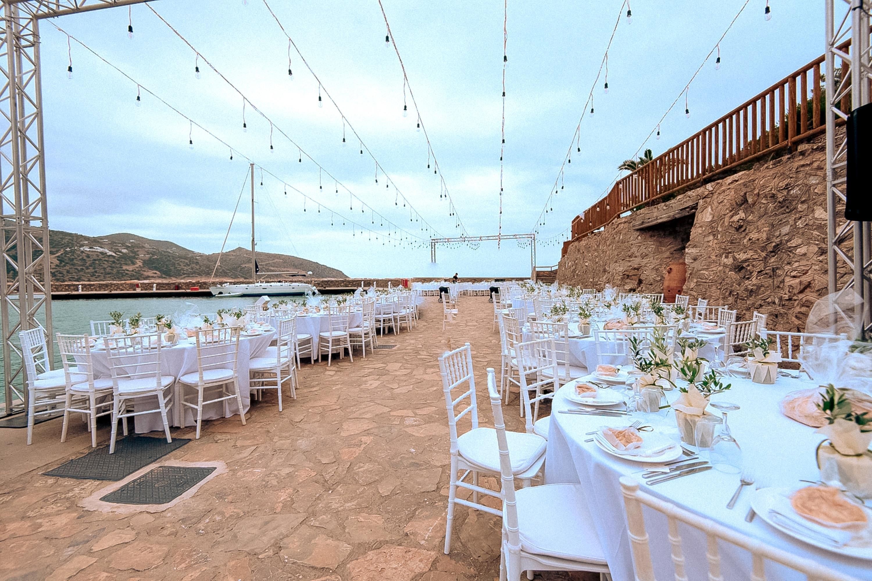 An outdoor dining area with white tables and chairs, set against a backdrop of a rocky cliff and a body of water.