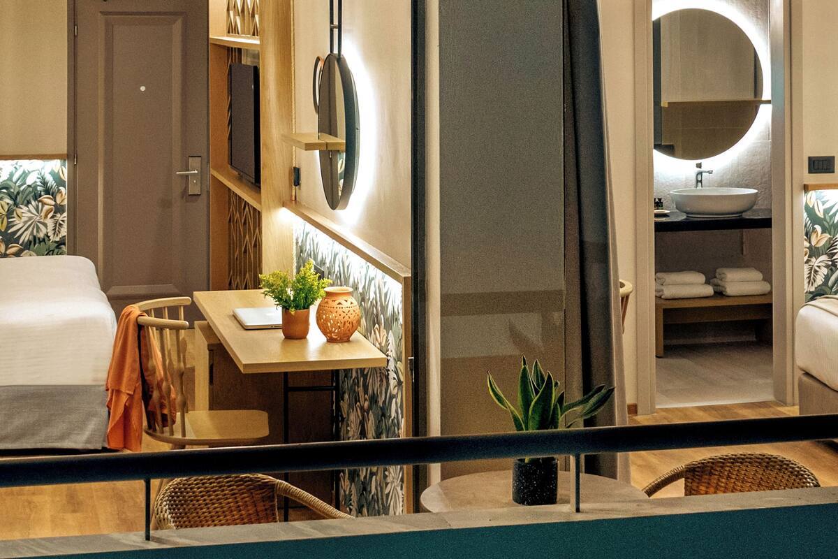 Una habitación de hotel con una cama, una mesa con una computadora portátil y una planta, un baño con un lavamanos y toallas, y vista al mar.