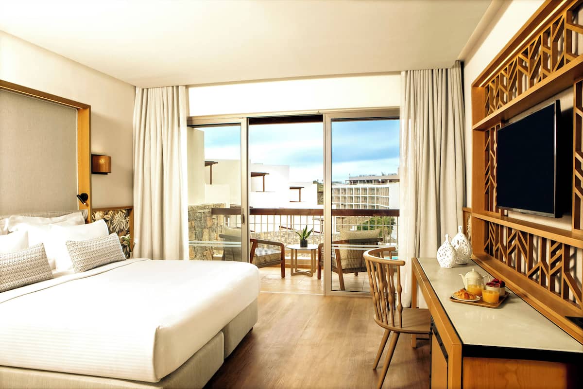 Habitación de hotel con una cama grande, un escritorio con televisor, un balcón con vistas a edificios y una zona de comedor con mesa y sillas.