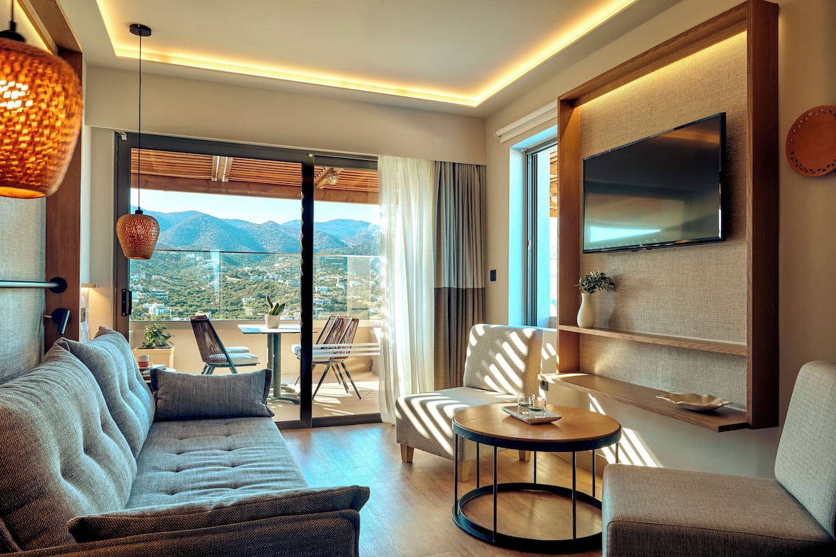 Una habitación de hotel moderna con sofá, sillón y una pequeña mesa. Un televisor está montado en la pared y hay un balcón con vistas a las montañas.