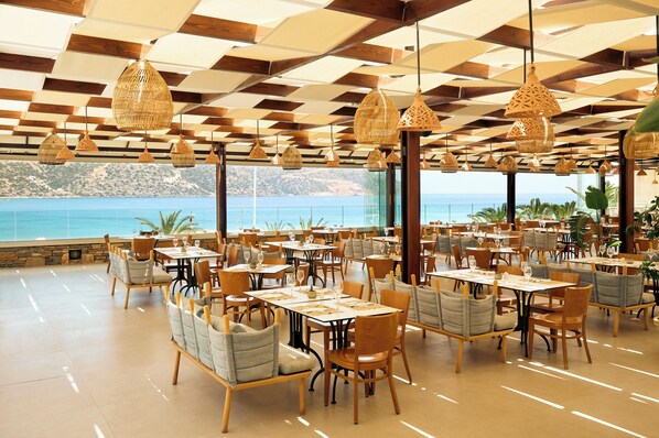 A spacious restaurant with multiple tables and chairs, large windows offering a view of the sea, and hanging wicker lamps. - Zeus Eleva Mirabello Bay (Agios Nikolaos)