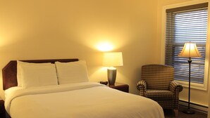 Ein Hotelzimmer mit einem großen Bett, einem Nachttisch mit Lampe, einem Sessel und einem Fenster mit Jalousien.