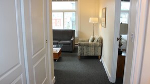 Ein schmaler Flur, der in ein Wohnzimmer mit grauem Sofa, gemustertem Sessel und einer Lampe führt.