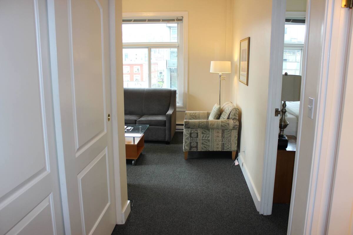 A narrow hallway leading to a living room with a grey sofa, patterned armchair, and a lamp.