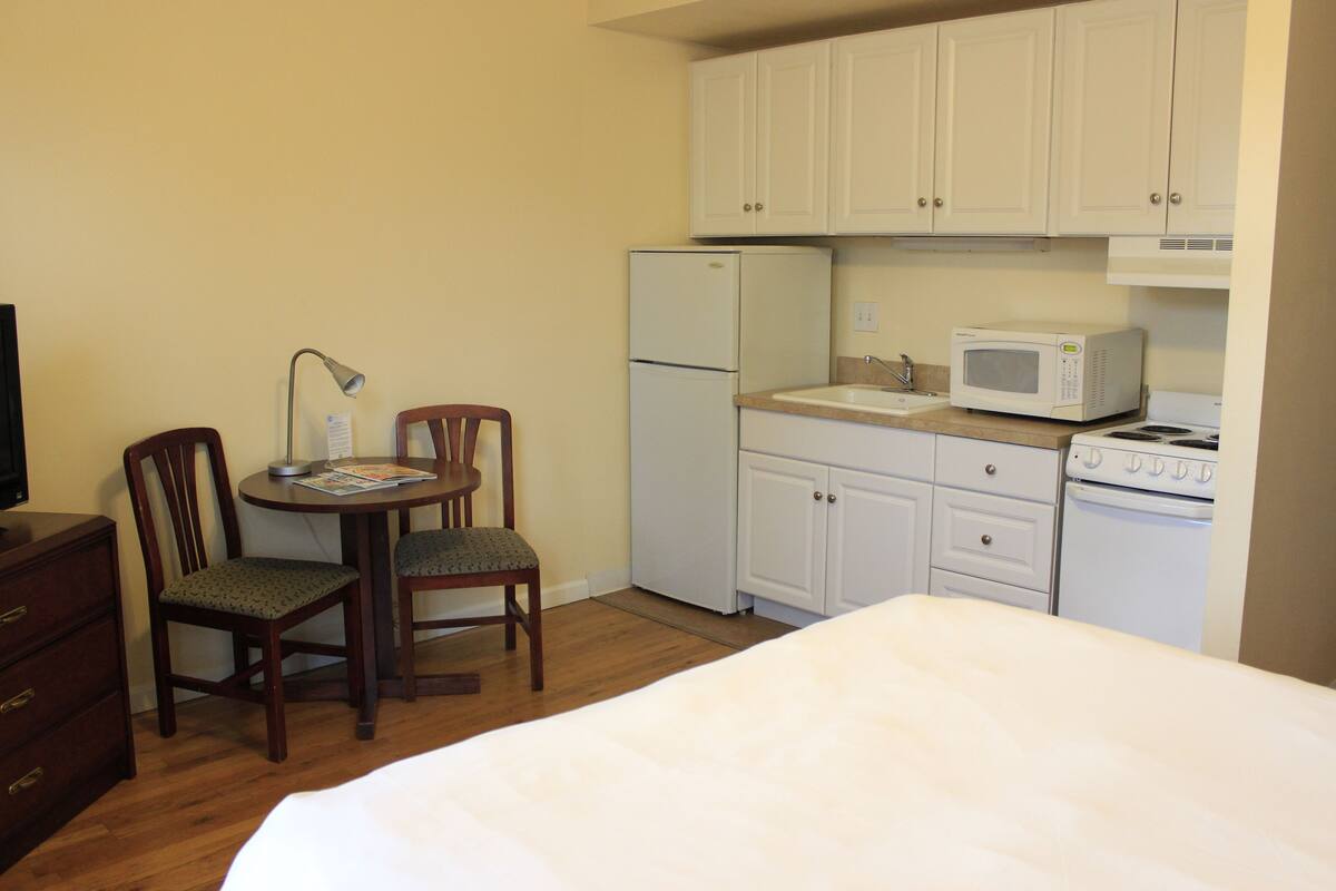 A compact hotel room with a bed, a desk with a chair, a small kitchenette with a refrigerator, microwave, and stove, and a television.