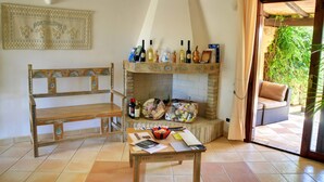 A cozy living room with a wooden bench, a table with papers, a fireplace with wine bottles, and a view of the outdoors. - Porto Giunco (Villasimius)