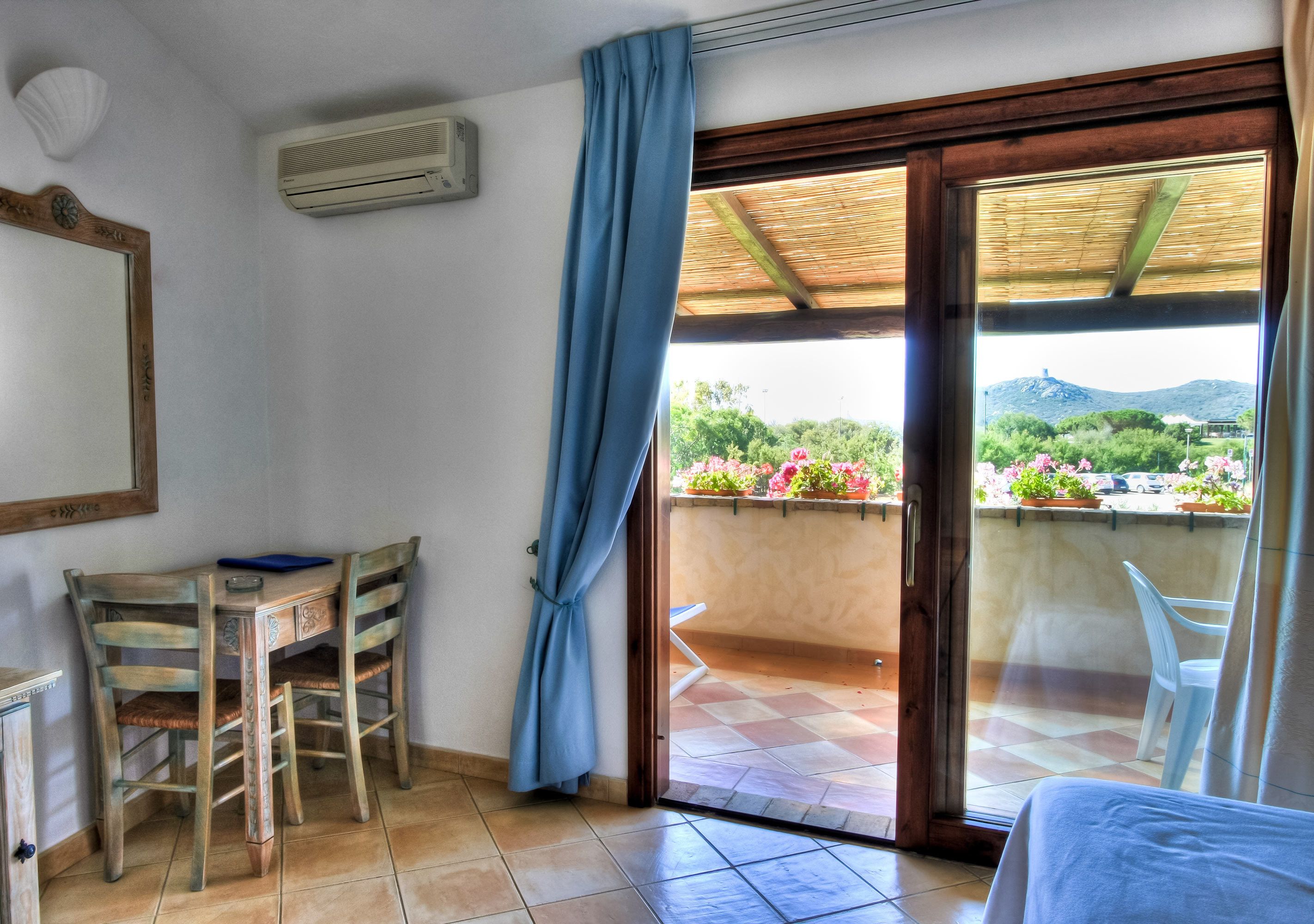 Una stanza con una scrivania in legno, due sedie, un letto e una porta scorrevole in vetro che dà su un balcone con vista su vegetazione e una montagna.