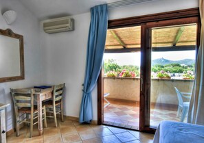 Habitación con escritorio de madera, dos sillas, una cama y una puerta corrediza de vidrio que da a un balcón con vistas a zonas verdes y una montaña.