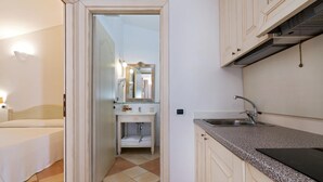 A compact kitchen with a sink, countertop, and cabinets. A mirror hangs above the sink, and a door leads to a bathroom with a bed and a toilet. - Porto Giunco (Villasimius)
