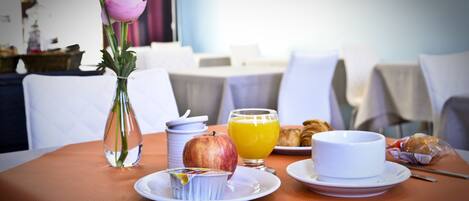 A breakfast table with a vase of flowers, a plate with an apple, a glass of orange juice, a cup, and a plate with croissants.