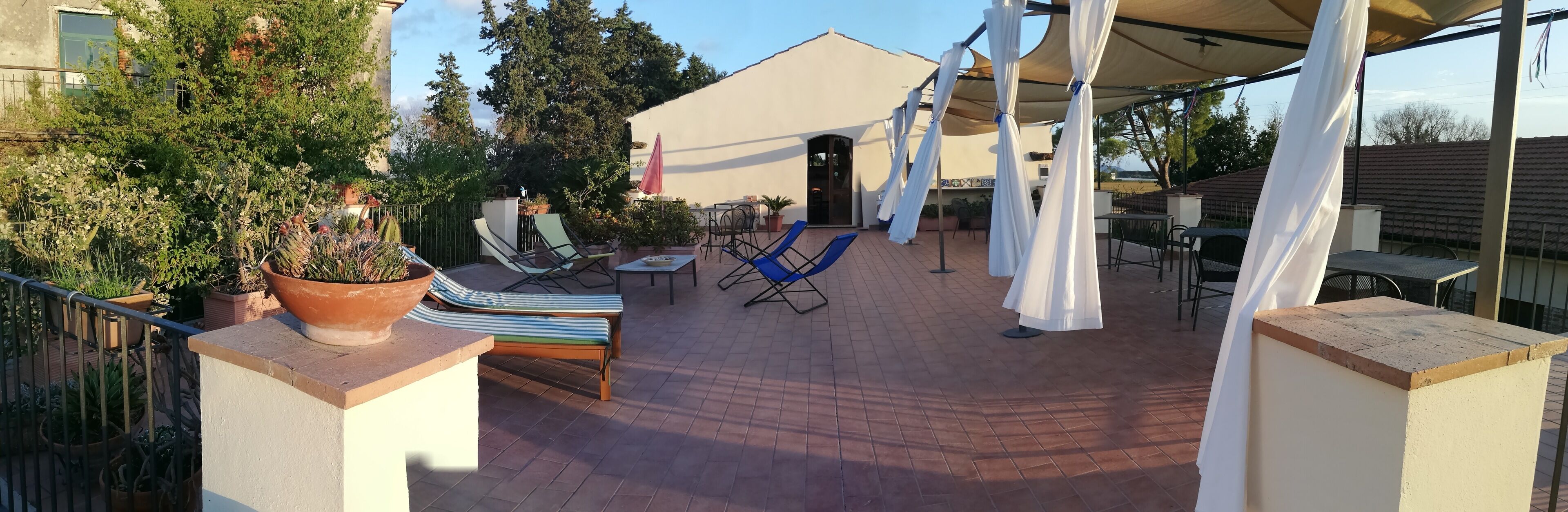 An outdoor patio with a covered seating area, potted plants, and a view of a building in the background.