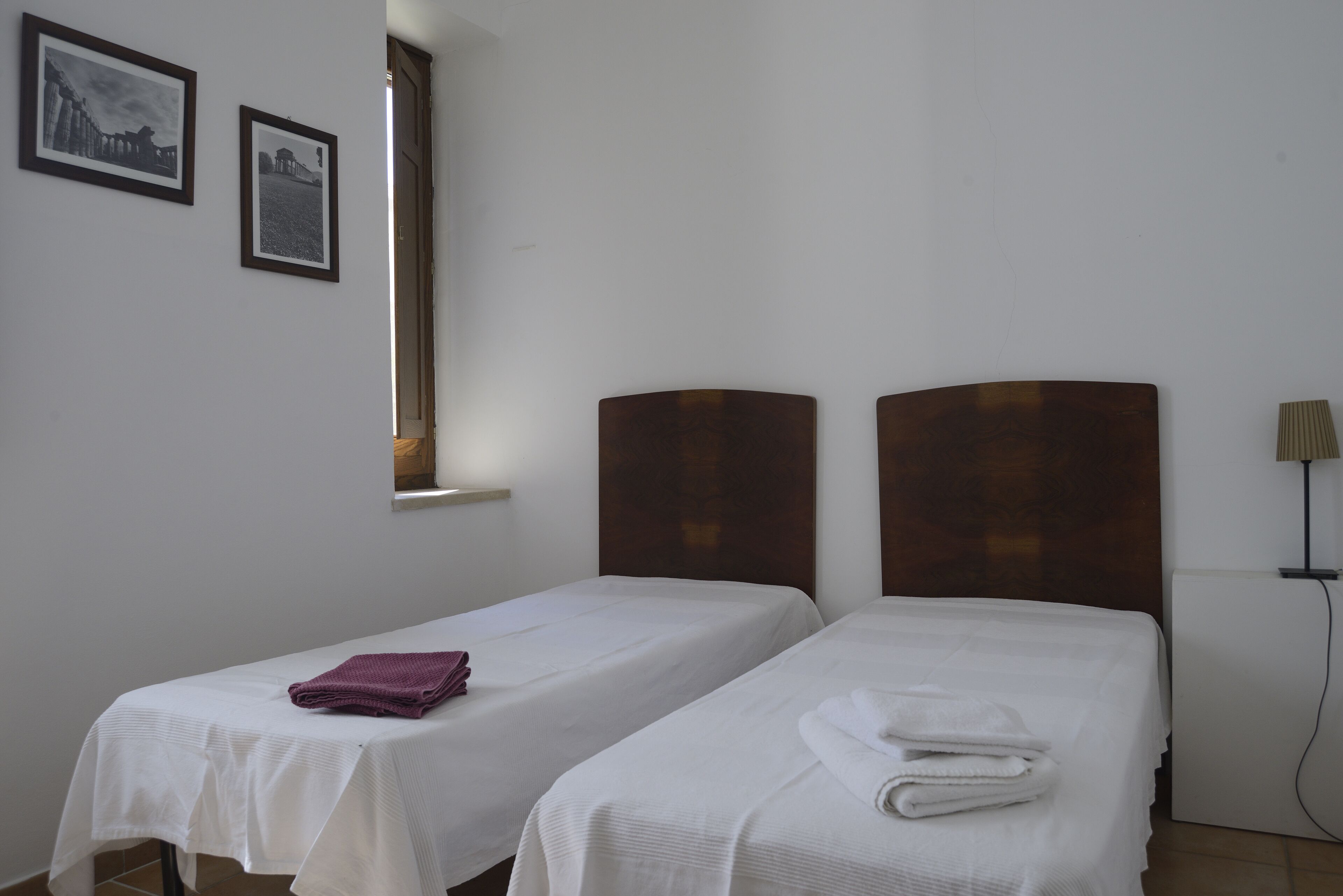 A hotel room with two single beds, a small table with a purple towel, and two framed pictures on the wall.
