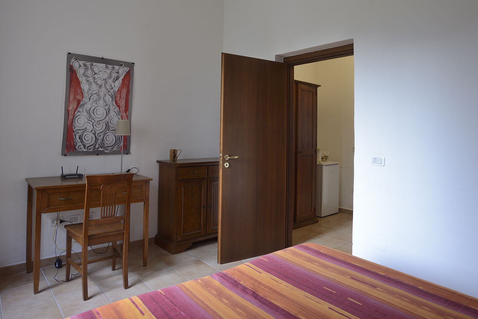 Una habitación con un escritorio de madera, una silla y un armario. Hay una puerta que conduce a otra habitación.