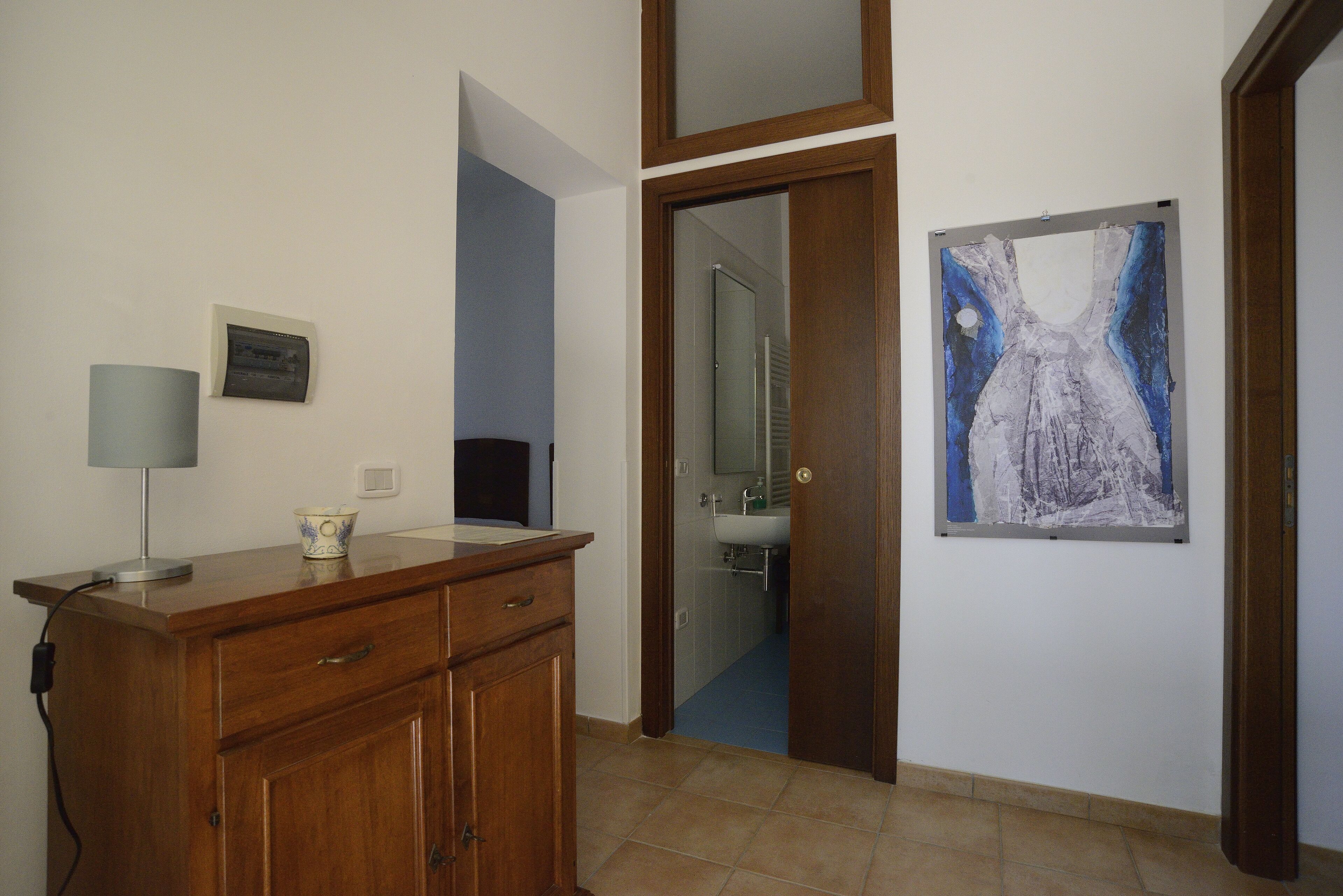A bathroom with a wooden cabinet, a lamp, a framed artwork, and a door leading to a shower area.