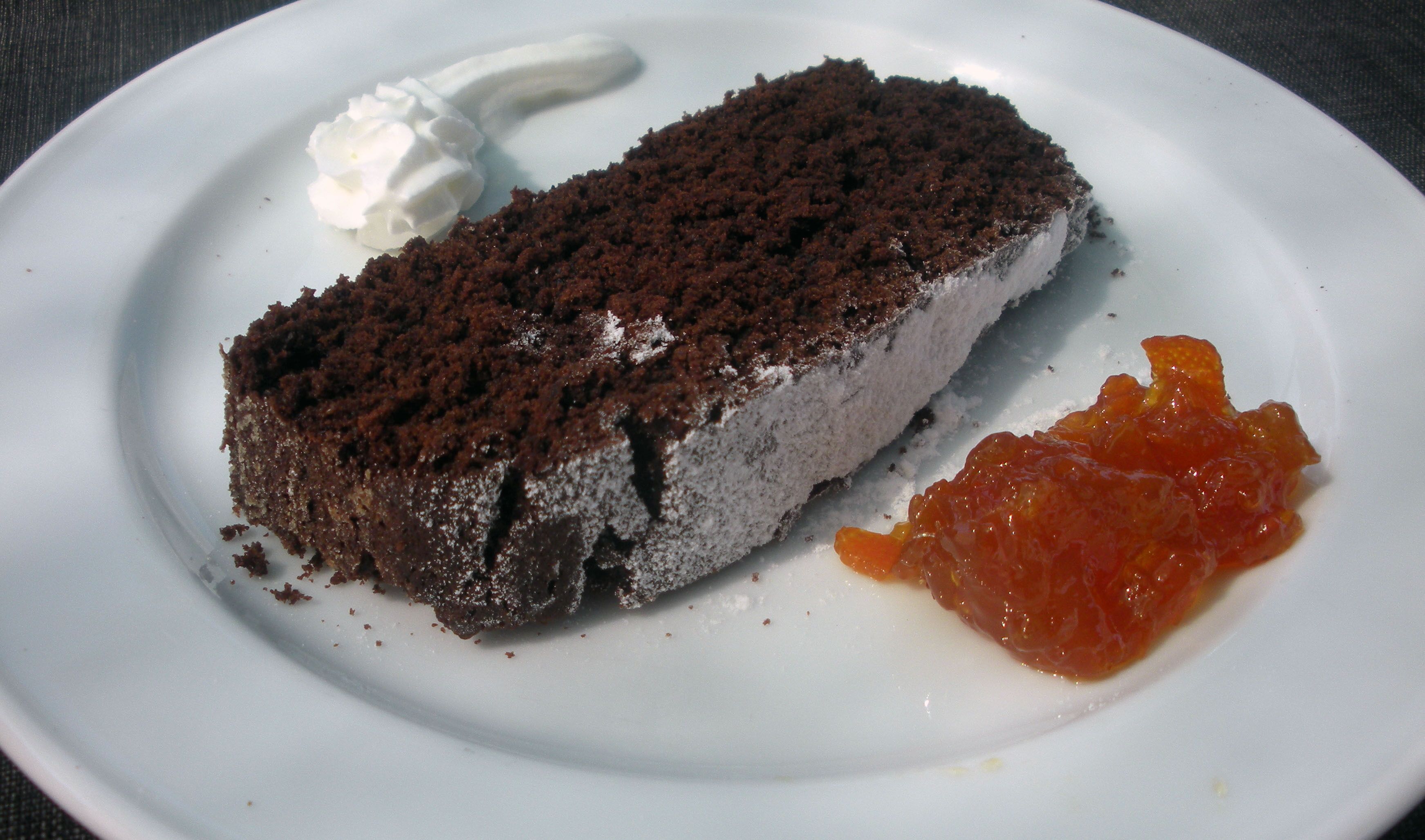 Une part de gâteau au chocolat, garnie de crème fouettée, accompagnée d’une portion de confiture de fruits, le tout présenté sur une assiette blanche.