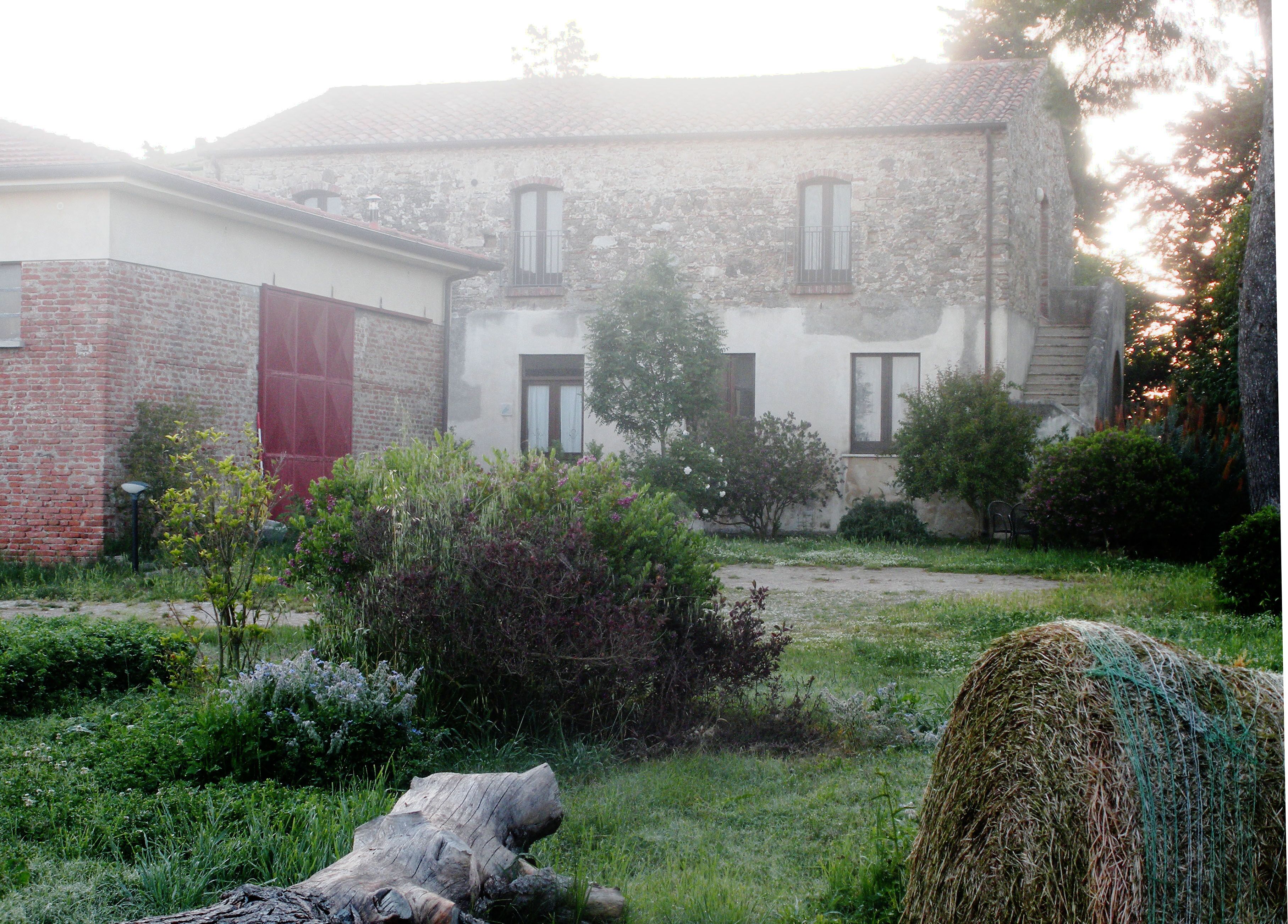 A rustic building with a brick facade, a red door, and a garden with hay bales and shrubs.