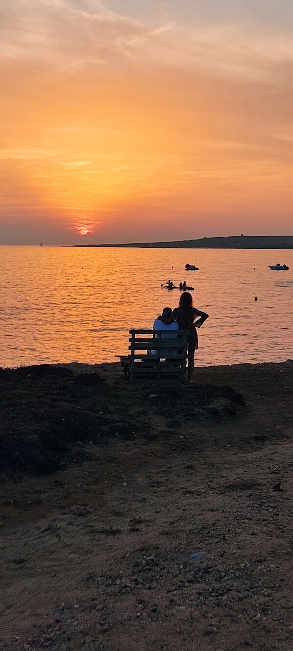 A sunset over a calm body of water with a wooden bench and people sitting on it. - B&B Italia (Milis)