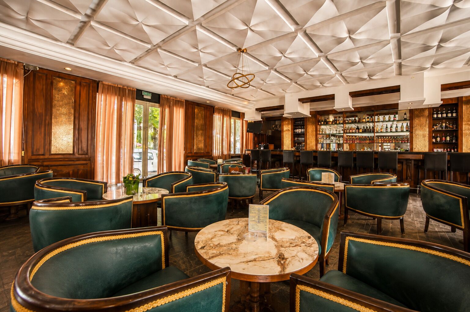 Zona de bar con sillas tapizadas de verde, una mesa de mármol y un techo con patrones geométricos.