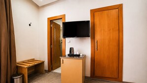 A hotel room with a wooden wardrobe, a television mounted on the wall, a small wooden desk with a coffee maker, and a trash bin. - Hotel Mamora (Kenitra)