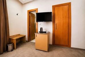 A hotel room with a wooden wardrobe, a television mounted on the wall, a small wooden desk with a coffee maker, and a trash bin. - Hotel Mamora (Kenitra)