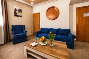Ein Zimmer mit einem blauen Sofa, einem blauen Sessel, einem Holztisch mit Früchten und einer Vase sowie einem runden, an der Wand befestigten Spiegel.