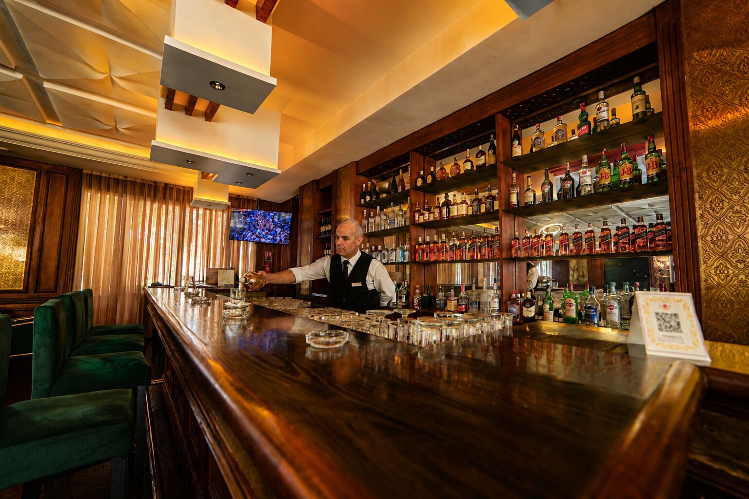 Zona de bar con un cantinero, estantes llenos de botellas de licor y taburetes.