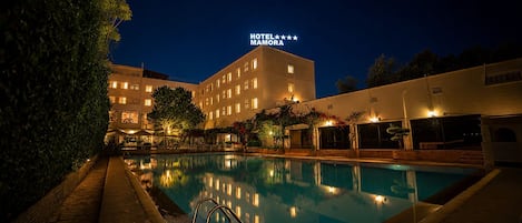 A hotel with a swimming pool at night, surrounded by trees and a well-lit building.