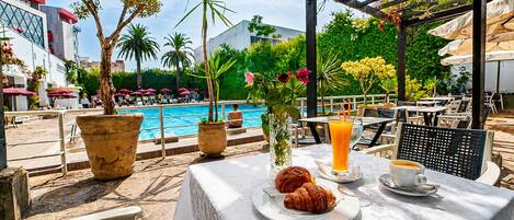 Un área de comedor al aire libre con piscina, plantas en macetas y sombrillas.